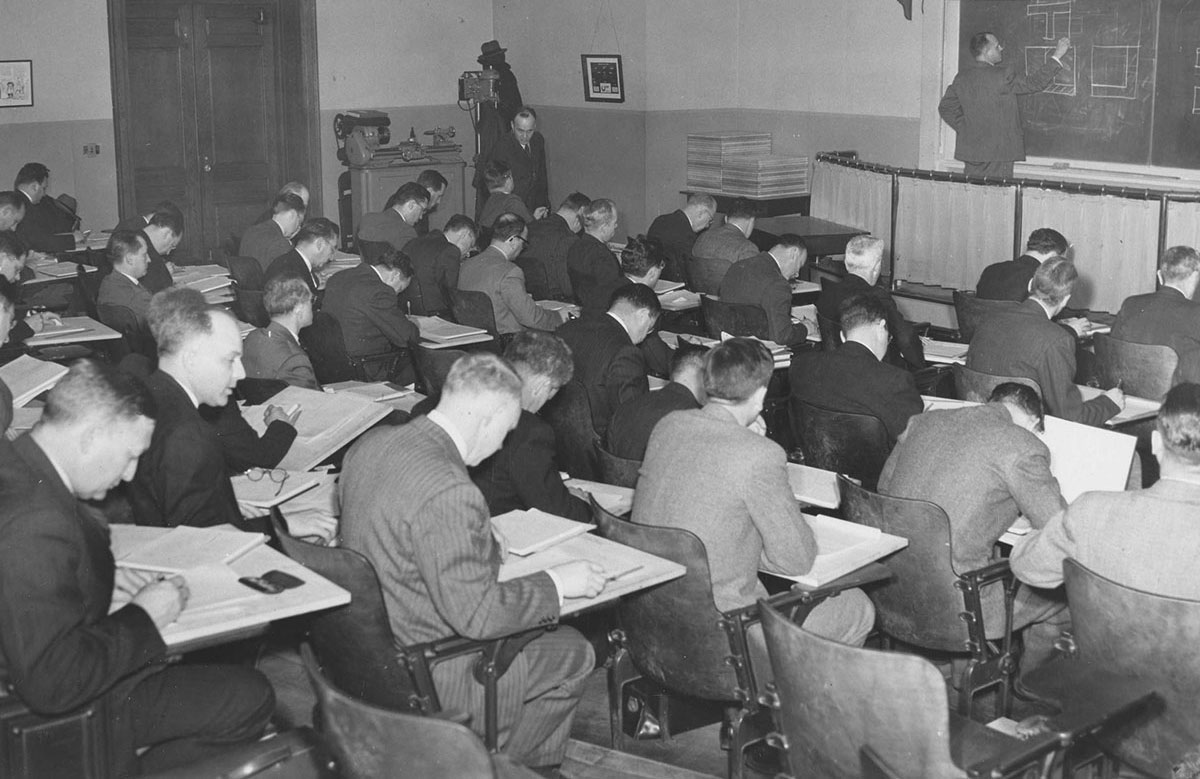 Black and white photograph of a classroom of men taking notes while a male professor writes on a chalkboard.