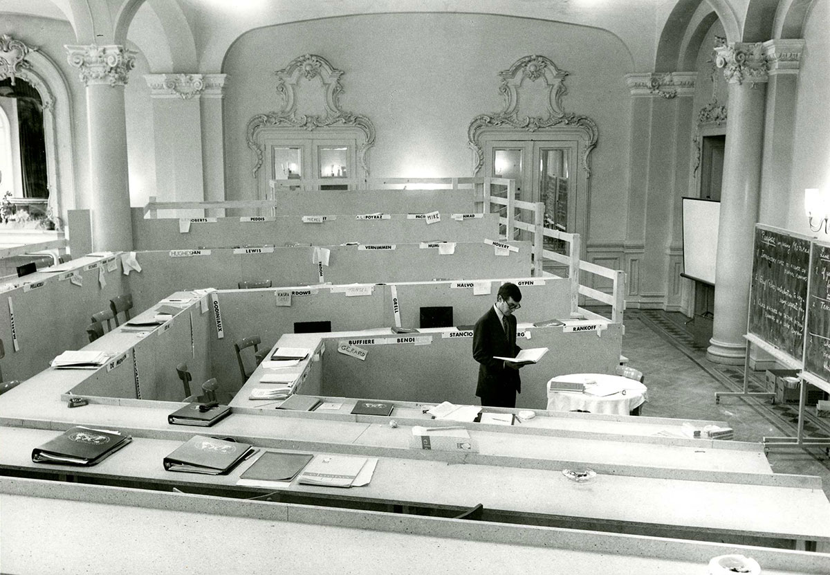 Black and white photograph of a man standing in the middle of an empty classroom, looking down at papers.