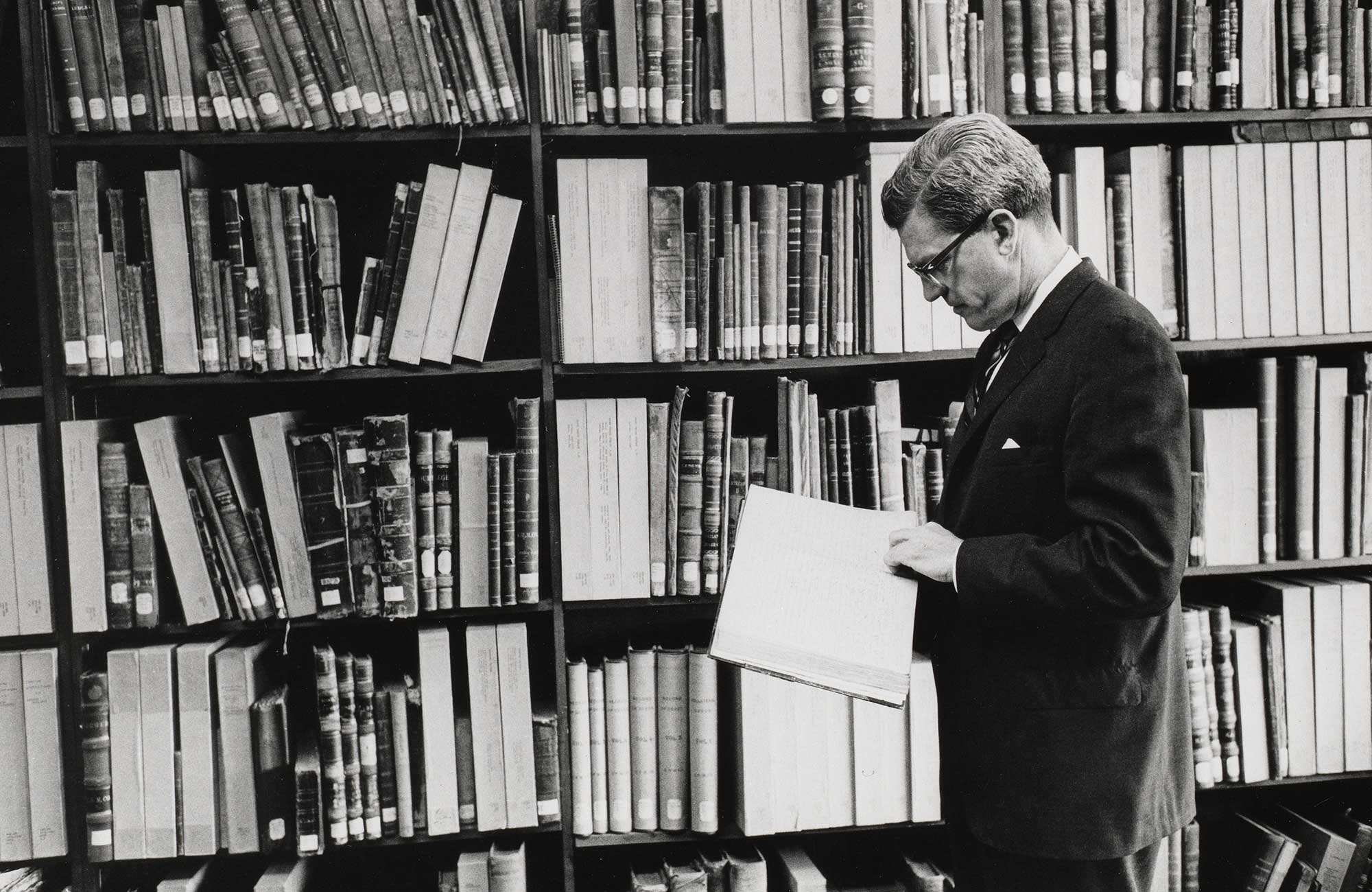 Black and white photograph of HBS Professor Kenneth Andrews reading a book in front of a bookshelf.