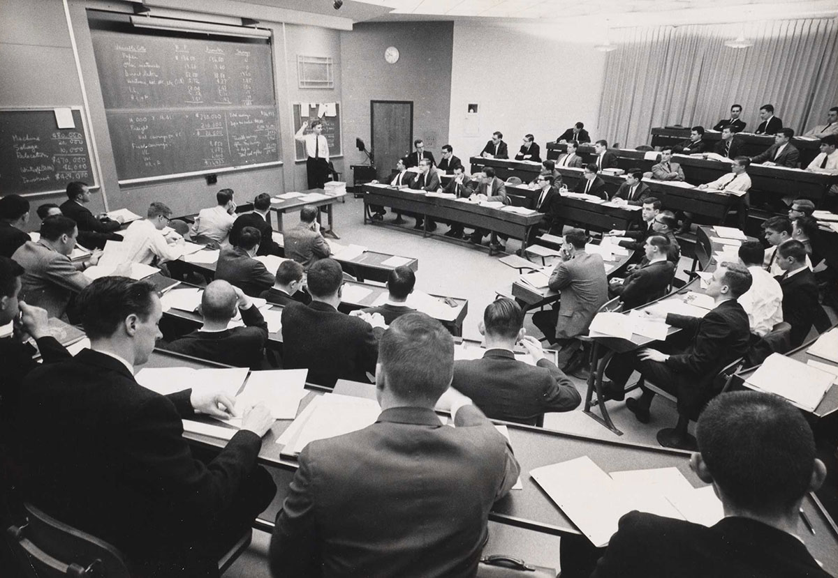 A classroom of male students in a tiered u-shaped classroom, looking toward a male professor gesturing near a chalkboard.