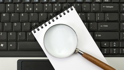 A laptop, a notebook, and a magnifying glass on a desk. (Shutterstock)